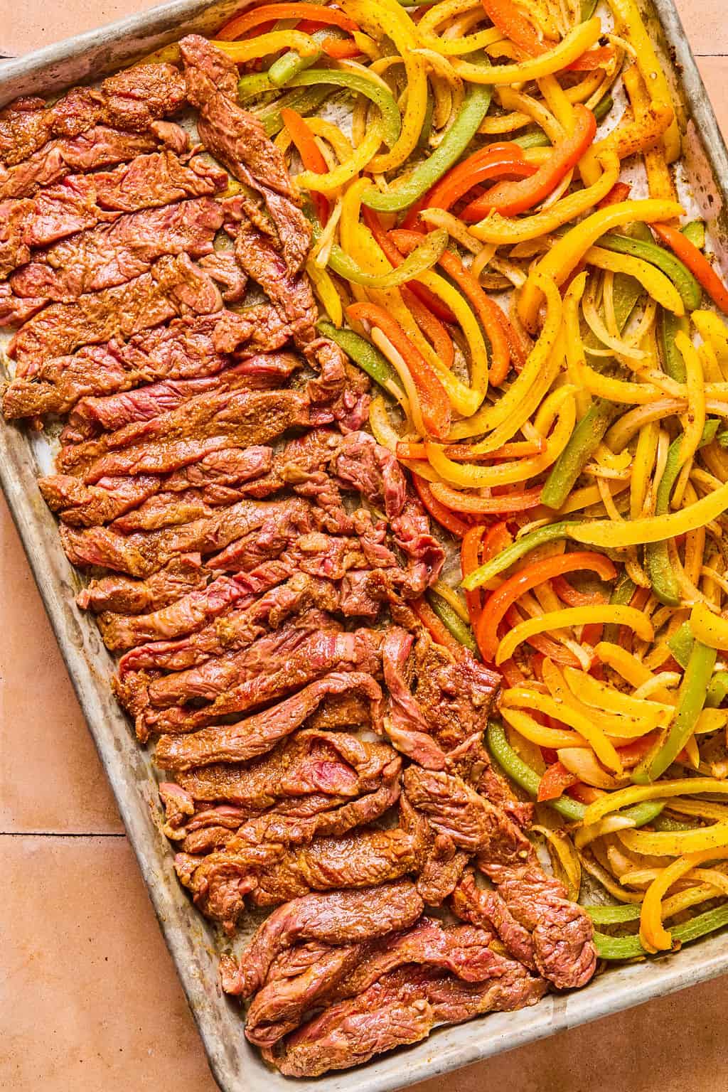 uncooked sliced flank steak fajitas and peppers and onions in a single layer on a sheet pan.