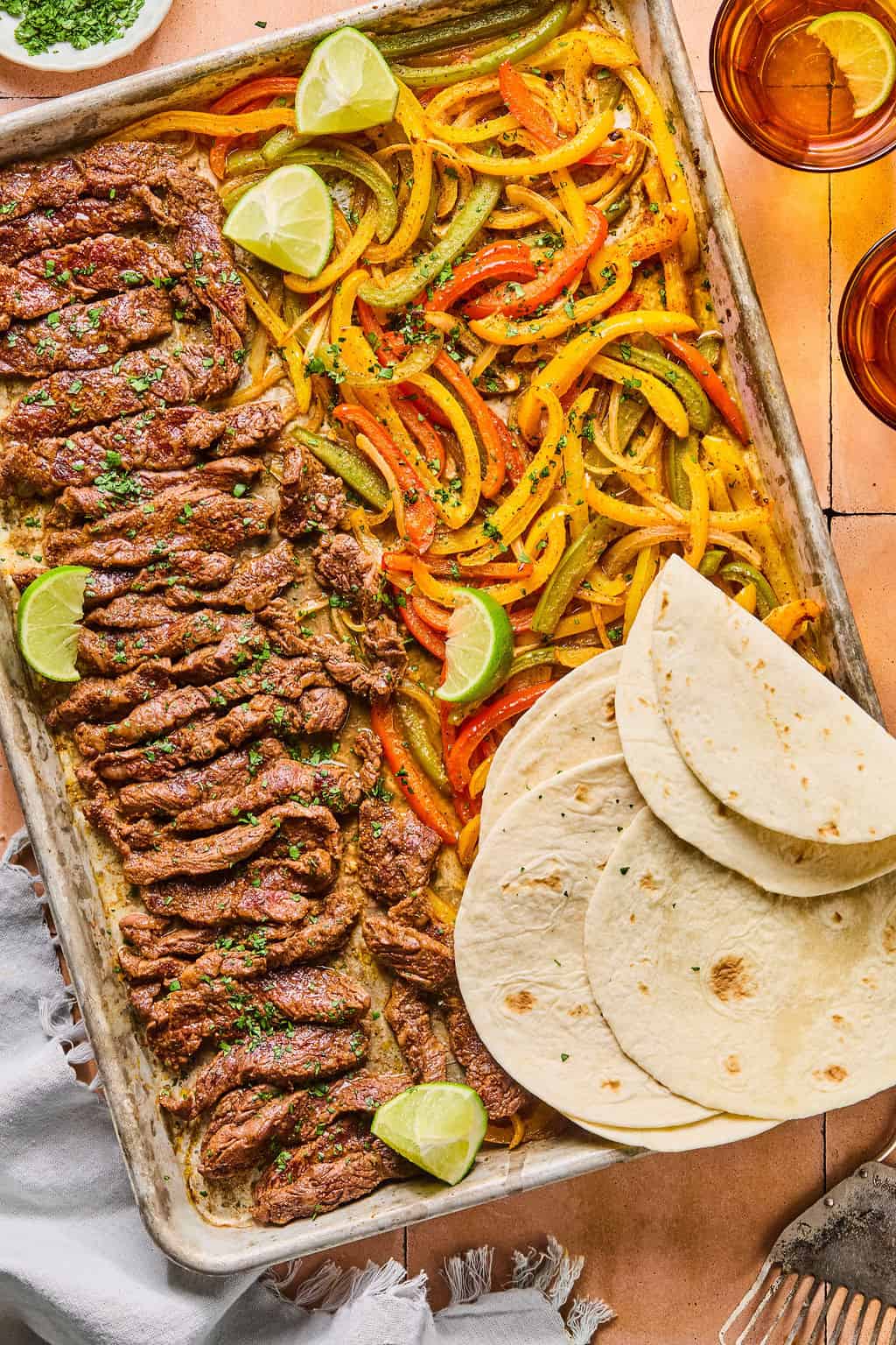 sheet pan steak fajitas with lime wedges and warm tortillas on the side.