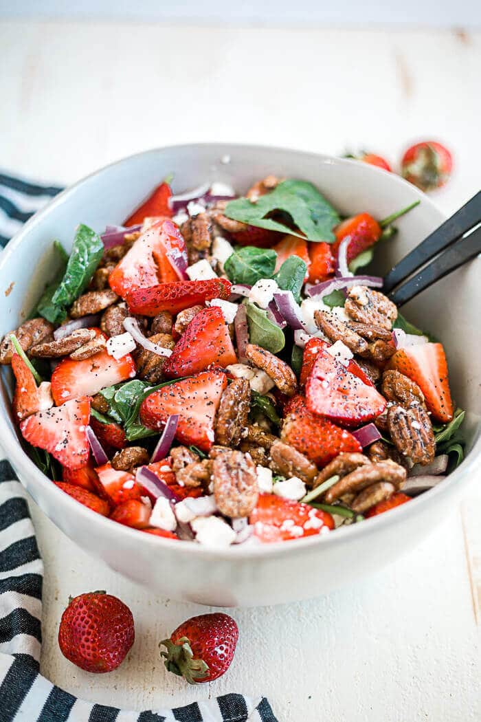 strawberry spinach salad coated in lemon poppyseed dressing in a large white serving bowl with serving utensils