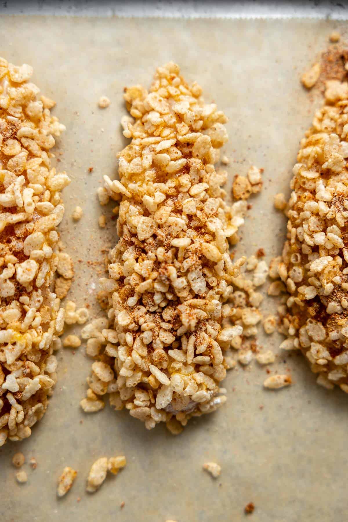 chicken tenders coated in rice krispie cereal on a baking sheet before cooking.