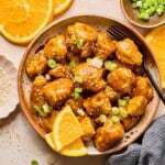 a bowl of crispy baked orange cauliflower in a bowl garnished with green onions and an orange wedge.