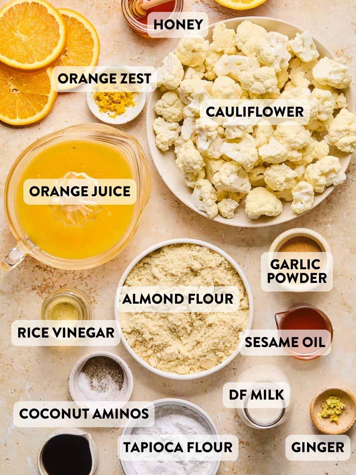 Overhead view of labeled ingredients for orange cauliflower, including fresh cauliflower florets, orange juice, orange zest, honey, almond flour, garlic powder, dairy-free milk, sesame oil, rice vinegar, coconut aminos, tapioca flour, black pepper, and fresh grated ginger.
