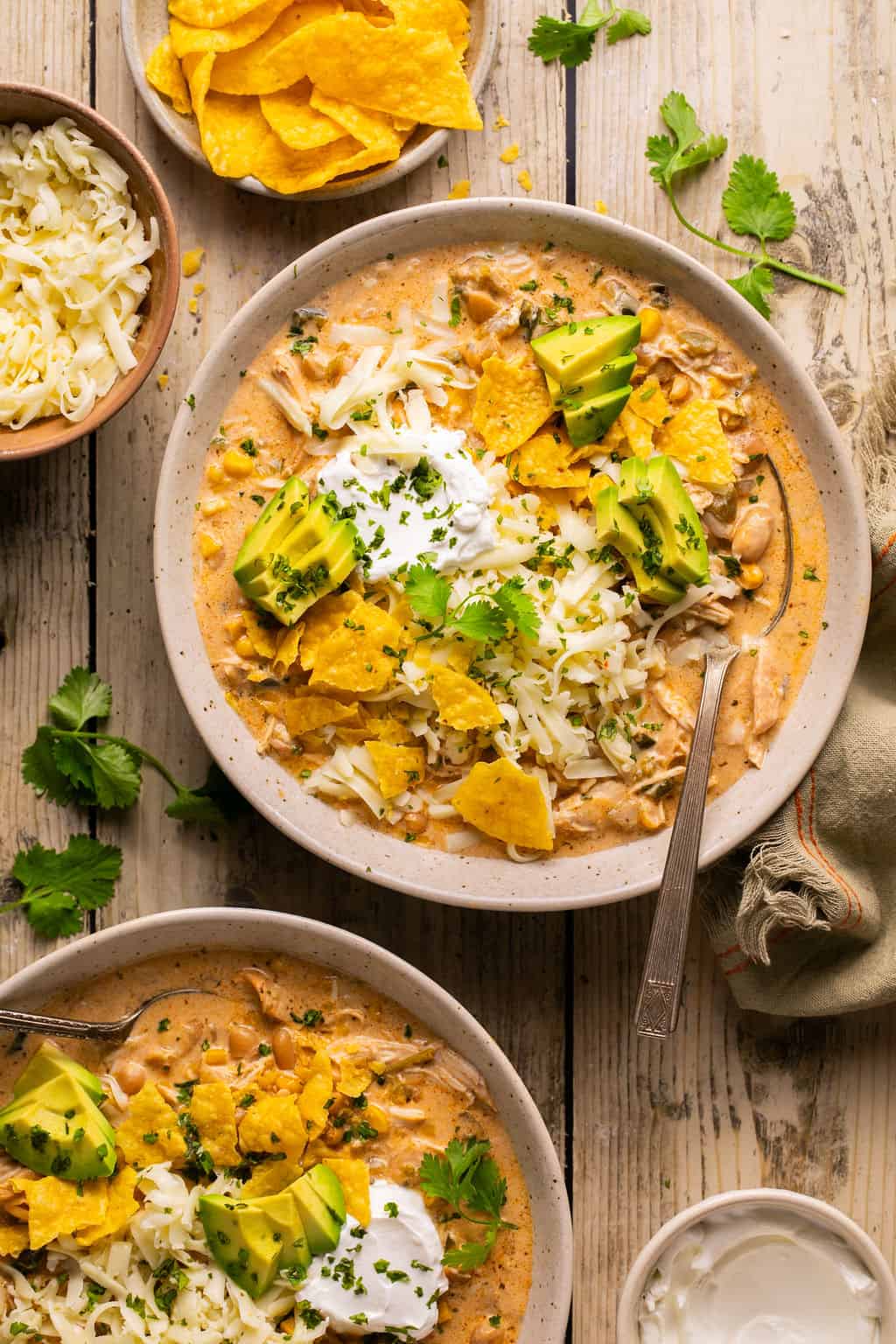 two bowls of healthy white chicken chili on a table with toppings around it.