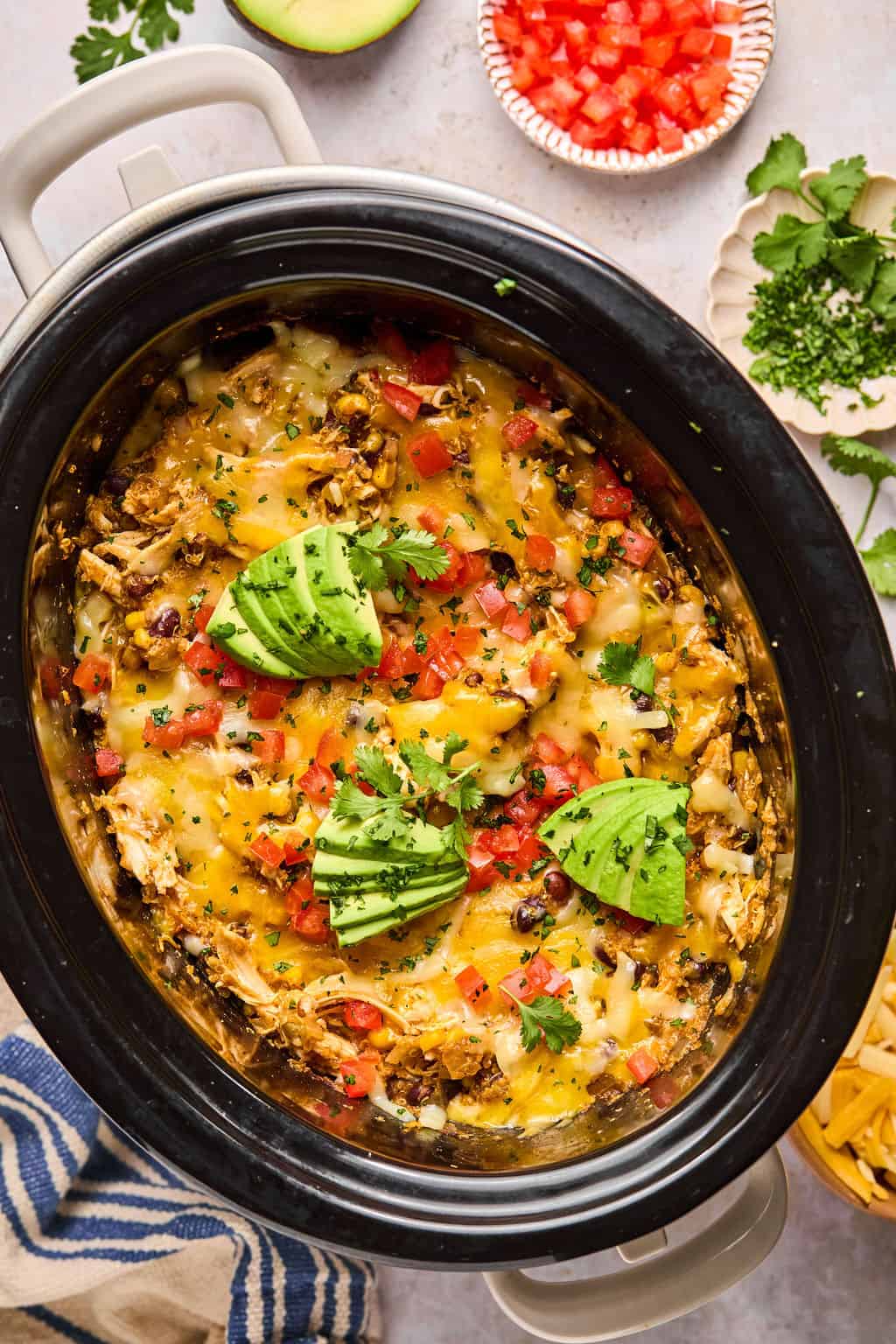 mexican casserole in a slow cooker topped with cheese, tomatoes, and sliced avocado.