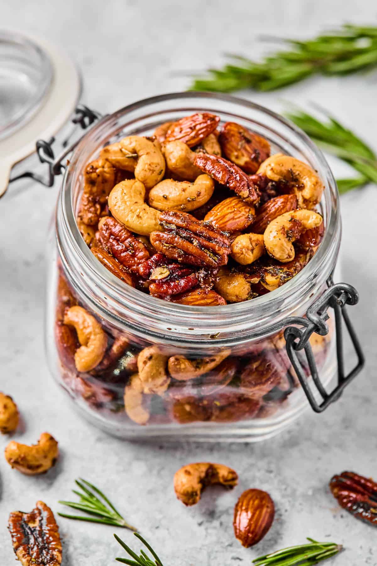 a jar of rosemary spiced nuts on a counter with fresh rosemary surrounding it.