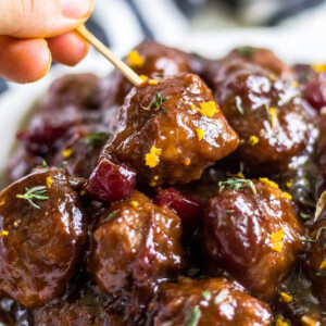 cranberry meatballs in a pile topped with orange zest and thyme with a toothpick picking one up