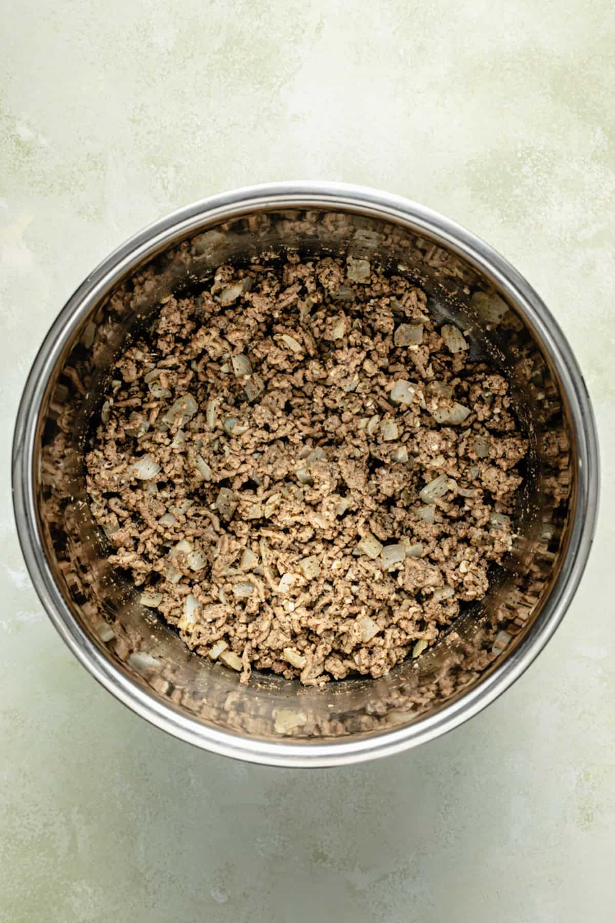 Cooked ground meat with chopped onions in a stainless steel bowl on a light textured surface.