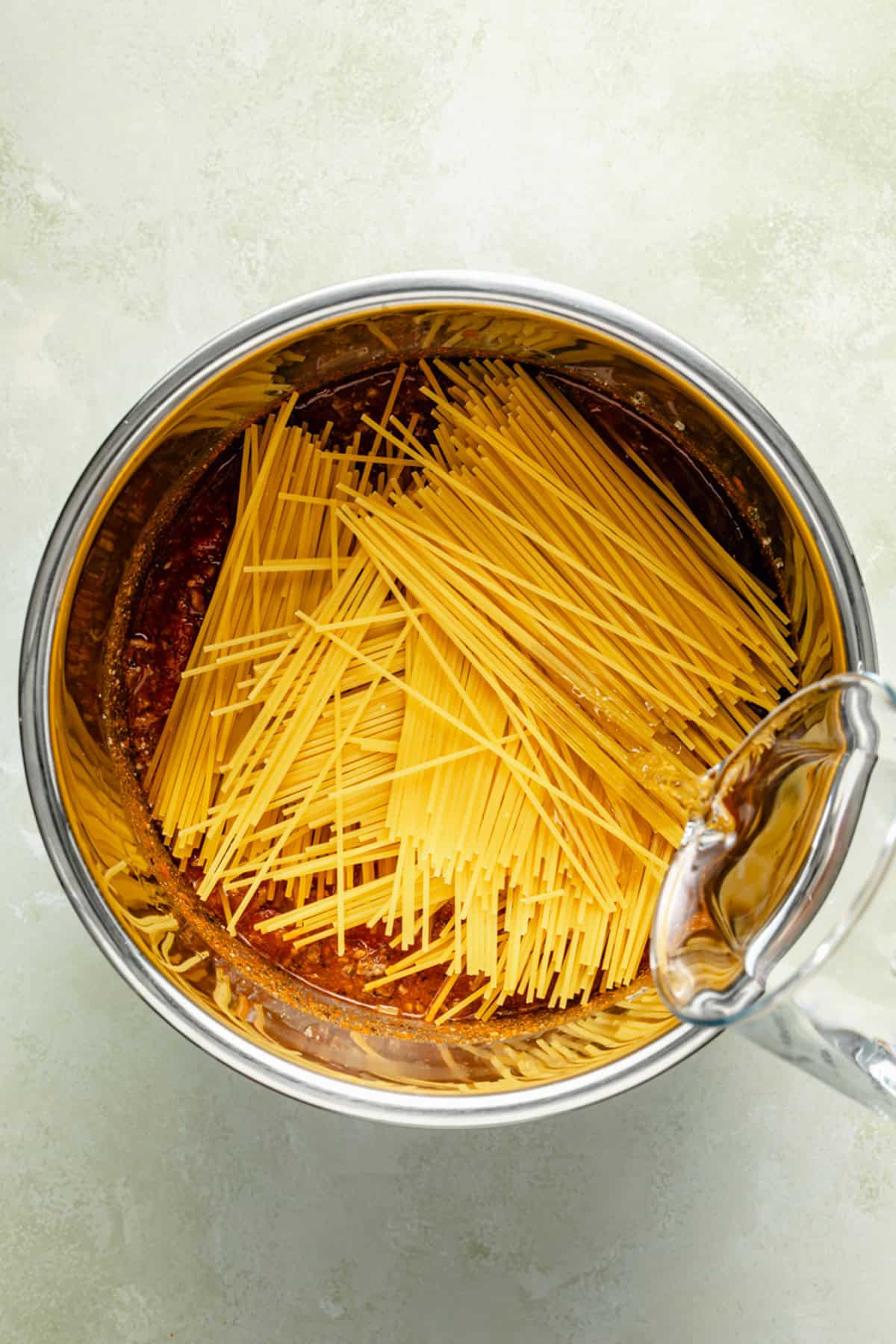 Uncooked spaghetti noodles in an isntant pot with tomato sauce, as water is being poured into the pot from a glass container—a simple start for instant pot spaghetti and meat sauce.