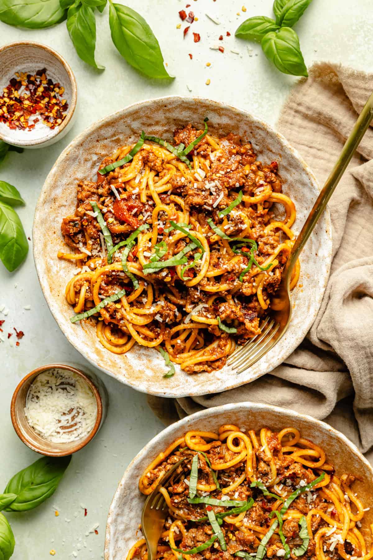 Two bowls of instant pot spaghetti and meat sauce, garnished with fresh basil and grated cheese. The bowls are on a beige cloth, surrounded by basil leaves and small bowls of grated cheese and red pepper flakes.