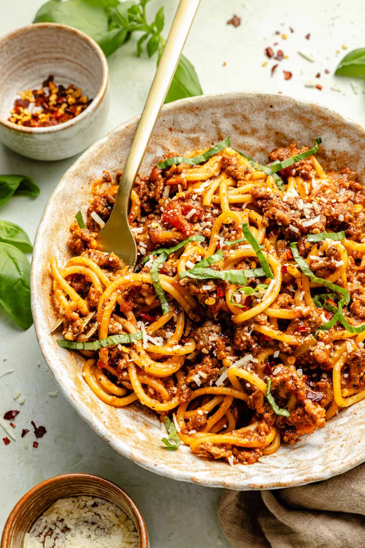 Bowl of spaghetti with meat sauce, garnished with chopped basil and grated cheese, with a fork in the bowl and small bowls of chili flakes and cheese nearby.