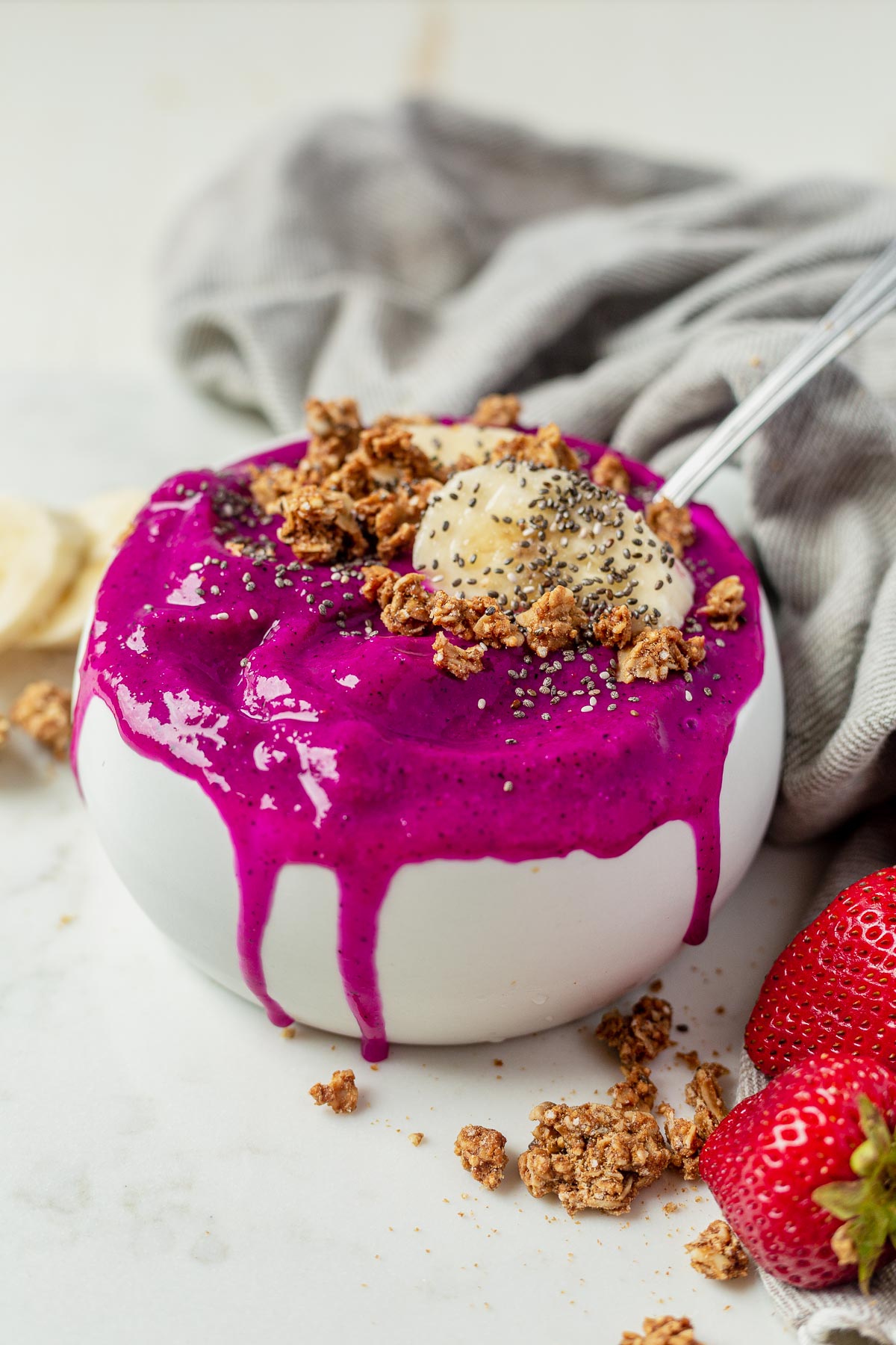bright magenta pitaya bowl dripping over the edge of a white bowl topped with granola and bananas