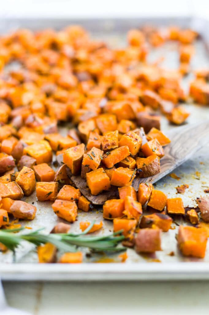 cubed and roasted sweet potatoes on a sheet pan with a wooden spoon and fresh rosemary