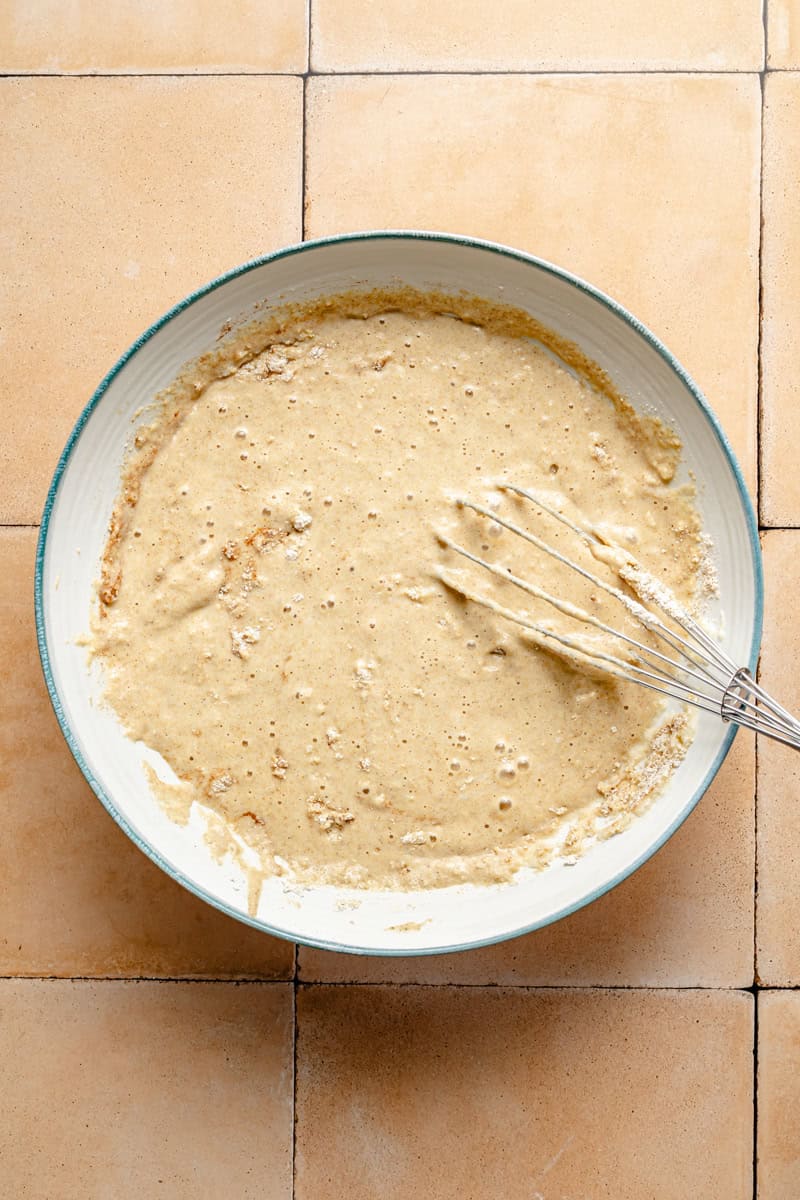 oatmeal waffle batter in a bowl on a pale pink tile counter with a whisk resting in the bowl.