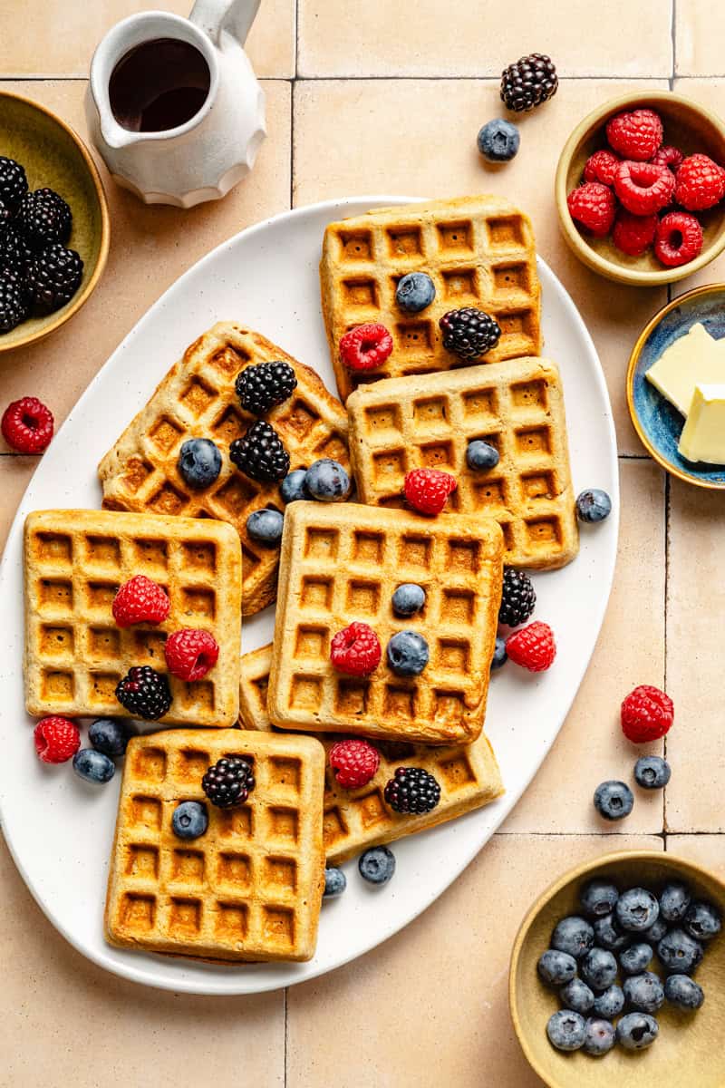 a platter of oatmeal waffles on a counter topped with fresh berries and a pitcher of maple syrup next to it.