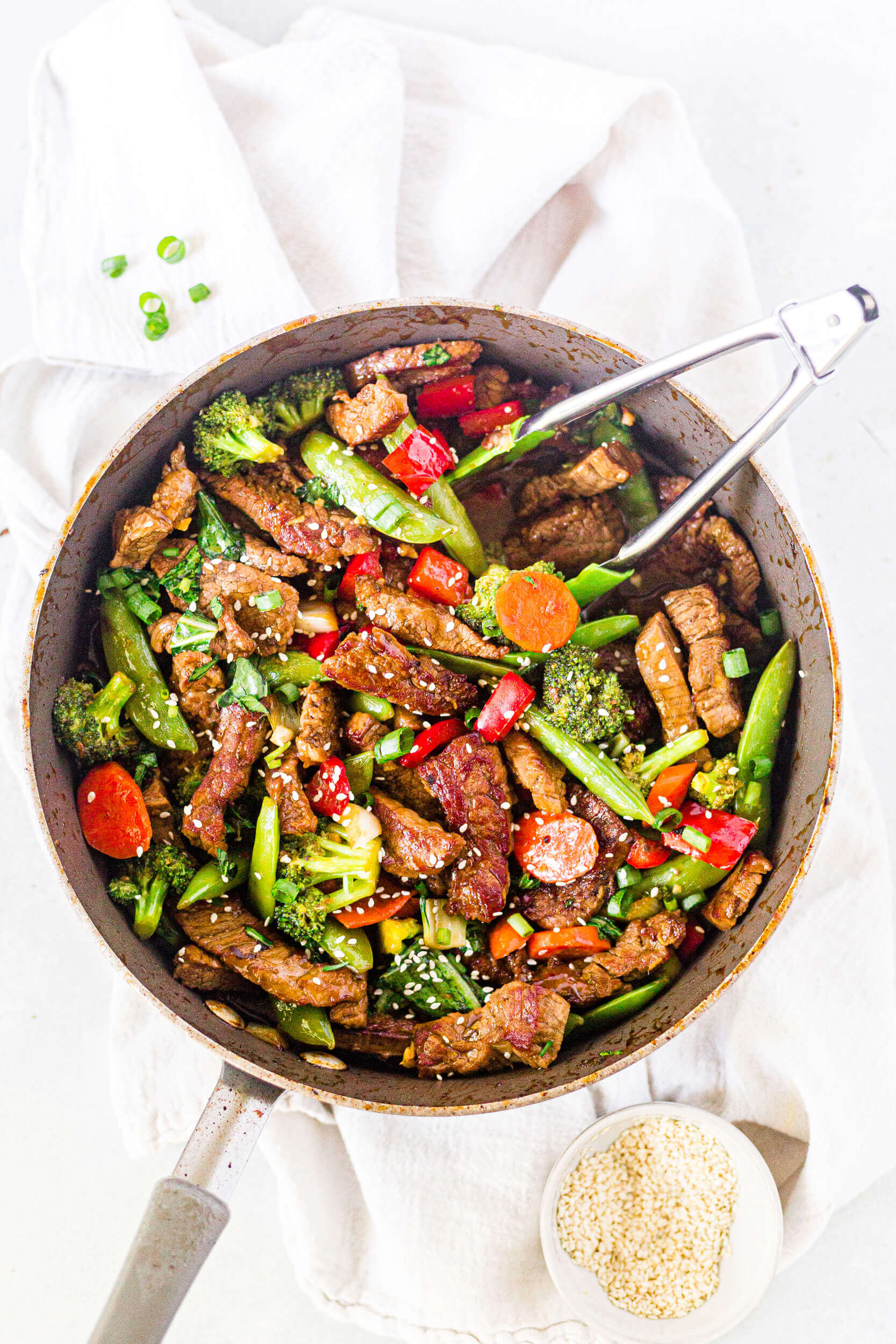veggie packed steak stir fry in a frying pan with tongs and sesame seeds