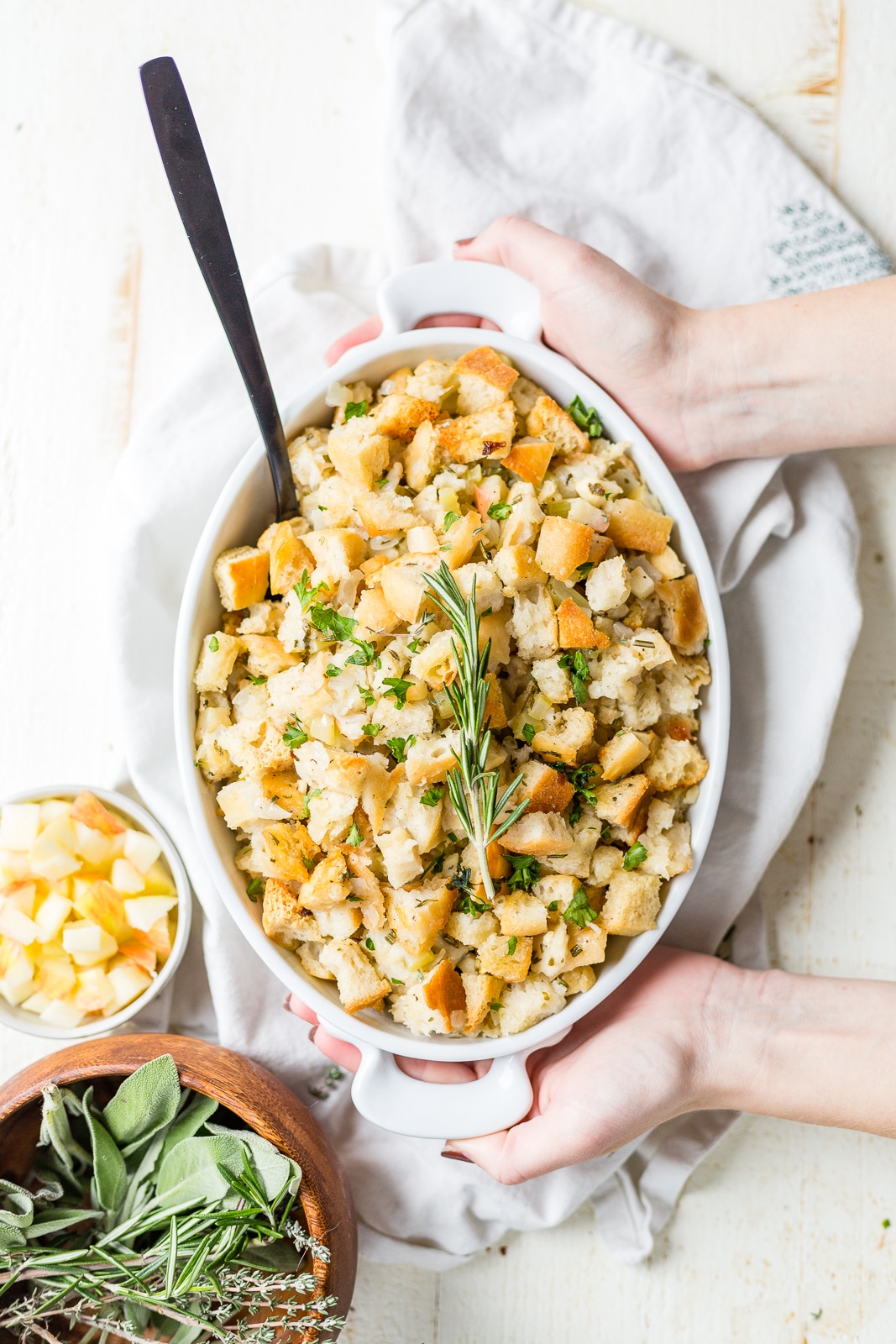 two hands picking up a dish of fresh simple sourdough stuffing topped with fresh herbs for thanksgiving