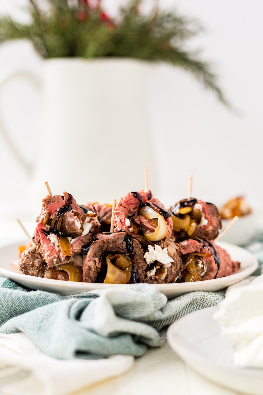 beef tenderloin roll up appetizers on a white plate with a green towel and christmas greenery in the back