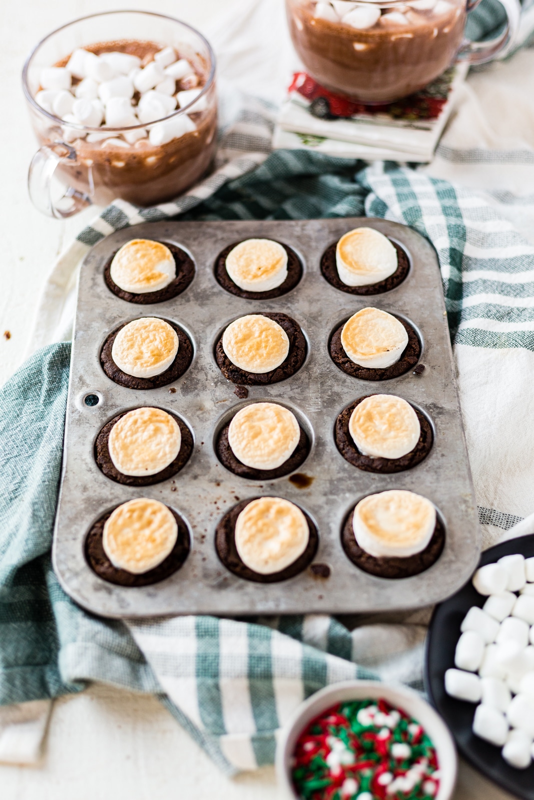 mini muffin tin holding baked hot cocoa cookies topped with a toasted marshmallow on a green towel