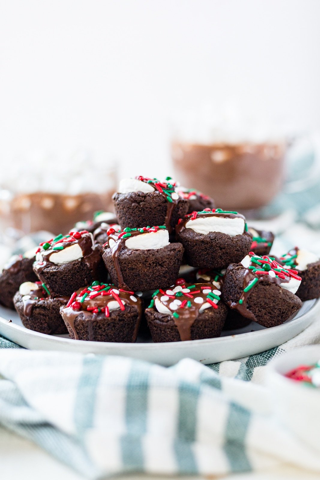 a platter of hot cocoa cookies topped with christmas sprinkles sitting on a green towel with warm hot cocoa in the background