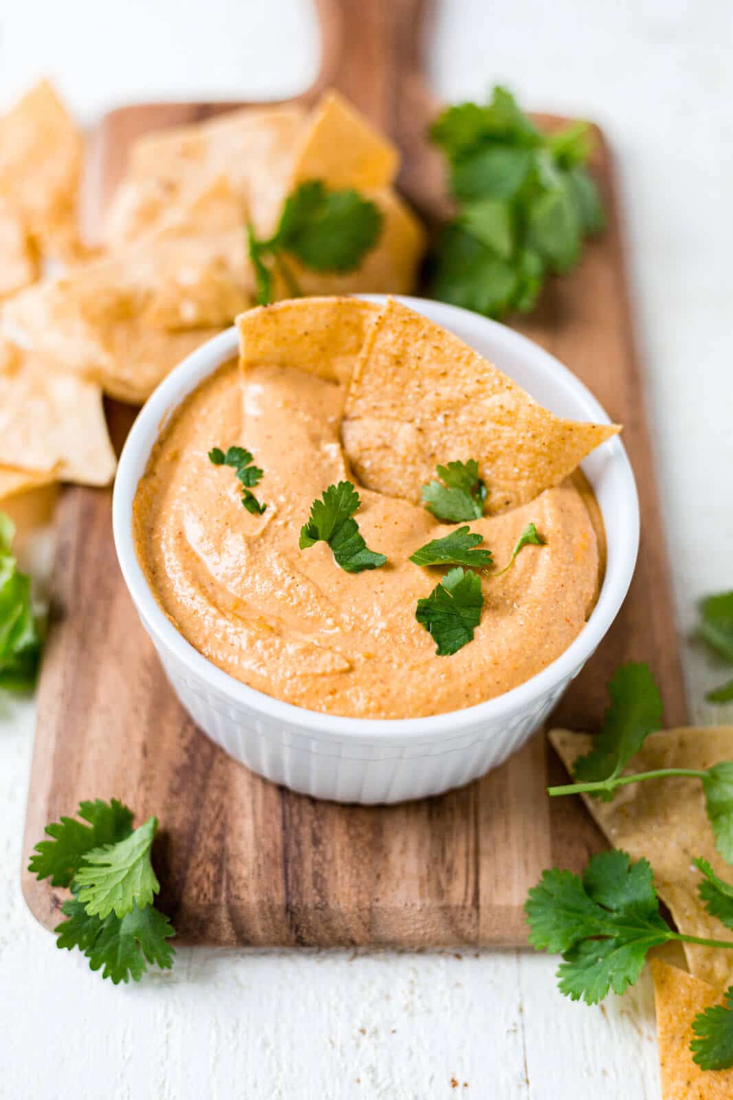 cashew queso in a white bowl sitting on a dark wood cutting board topped with chopped cilantro and tortilla chips