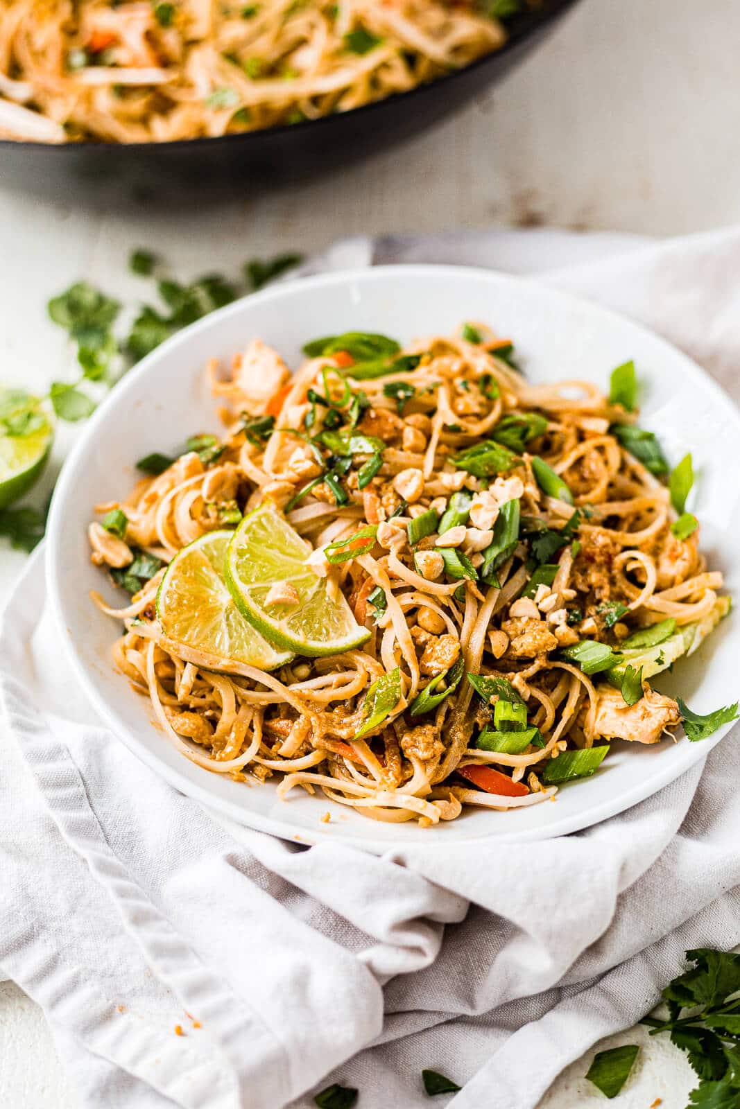 chicken pad thai topped with green onion, cilantro and lime in a shallow white bowl with a white napkin