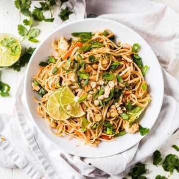 easy chicken pad thai recipe topped with green onion, cilantro and lime wedges in a shallow white bowl
