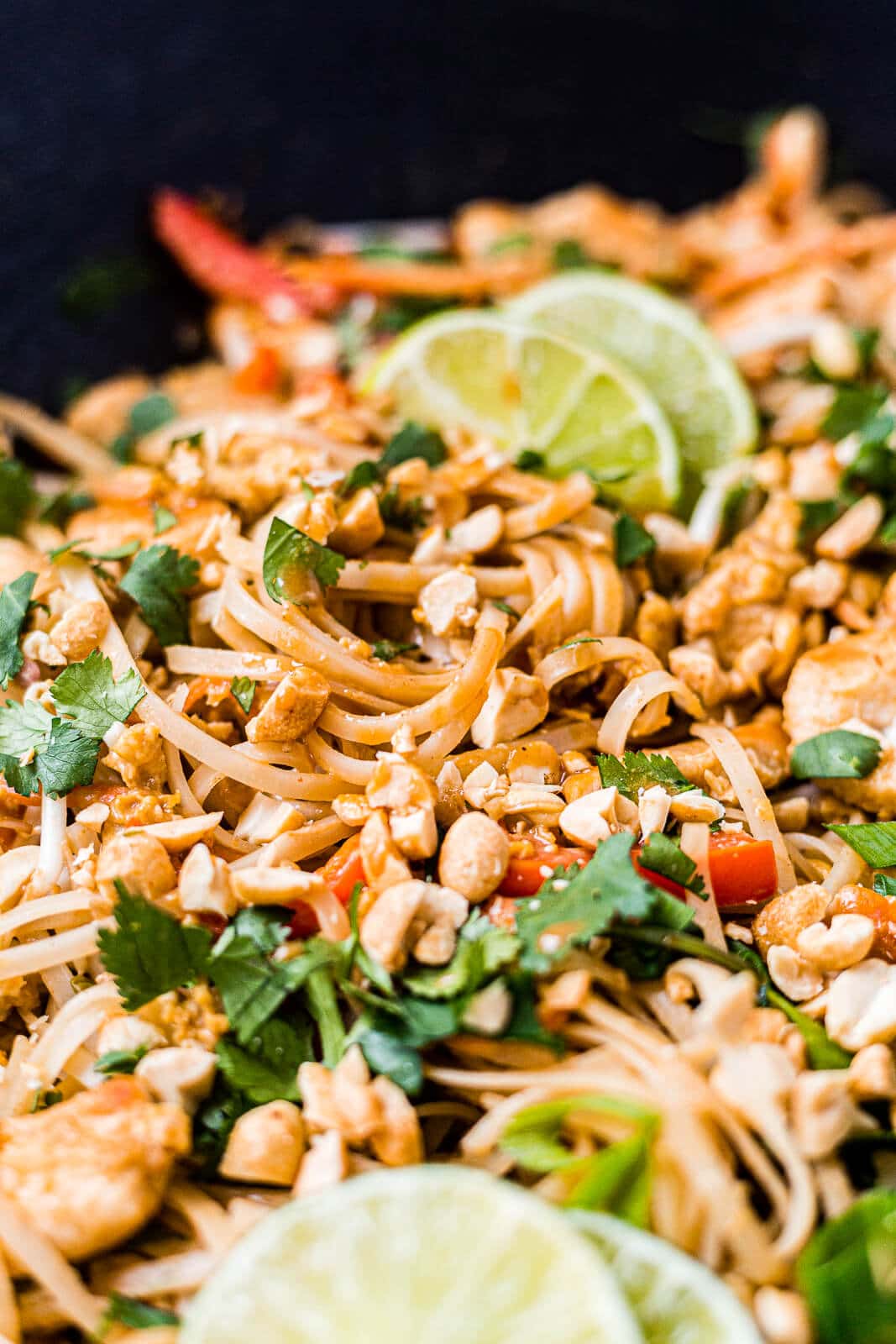 close up picture of chicken pad thai in a wok topped with cilantro, green onion and peanuts with lime wedges 