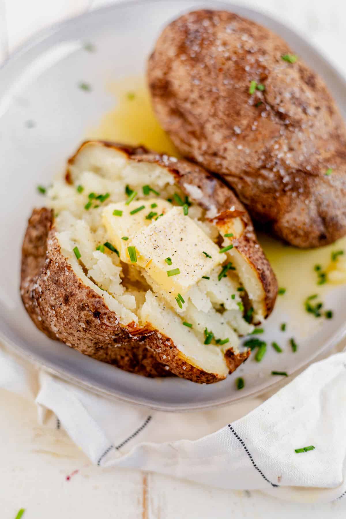 two air fryer baked potatoes on a plate topped with butter and chives.