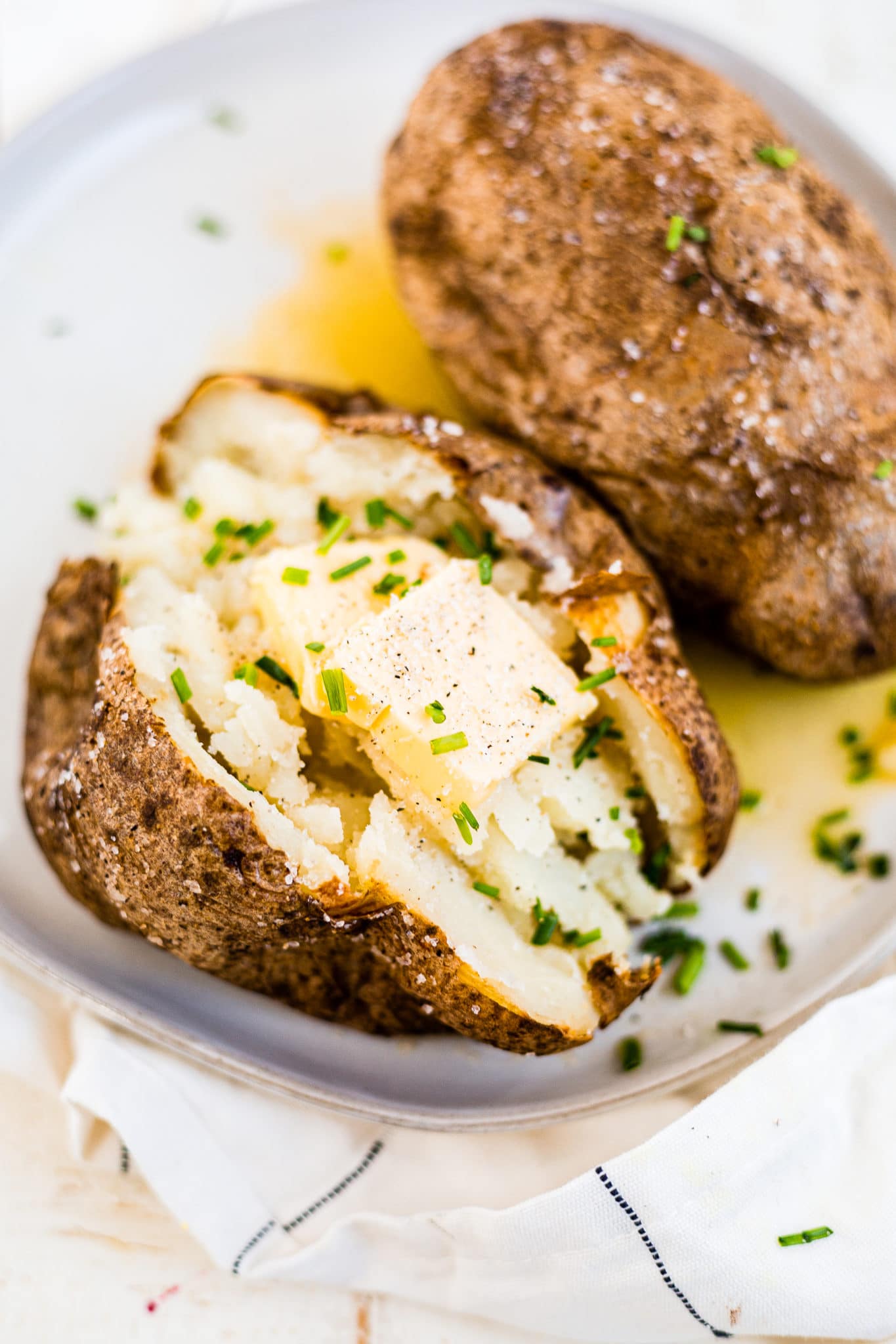two air fryer baked potatoes on a white plate topped with butter, salt, pepper and chives