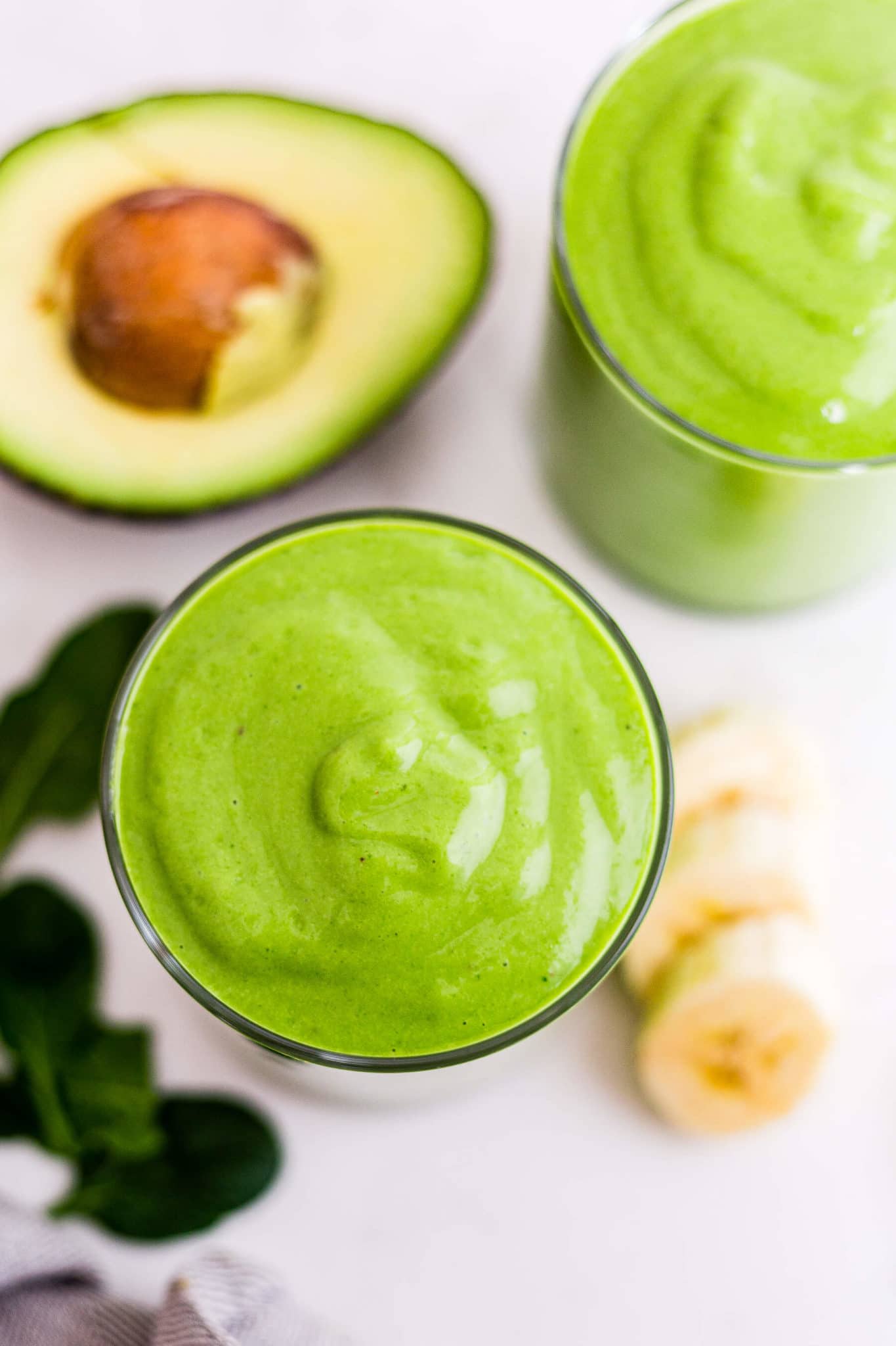 the top of a green smoothie in a glass with spinach, bananas and avocado around it  