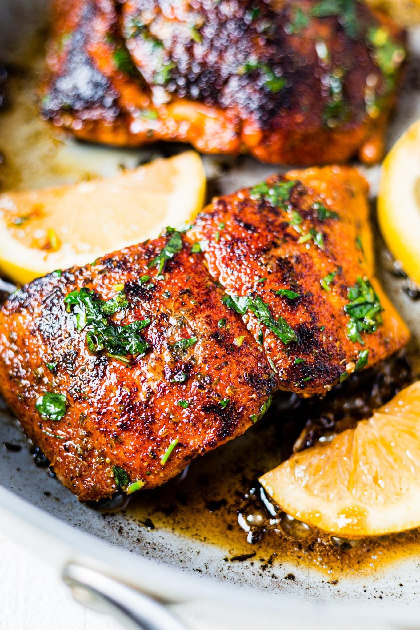 a piece of blackened salmon in a skillet with lemon wedges and topped with chopped parshley