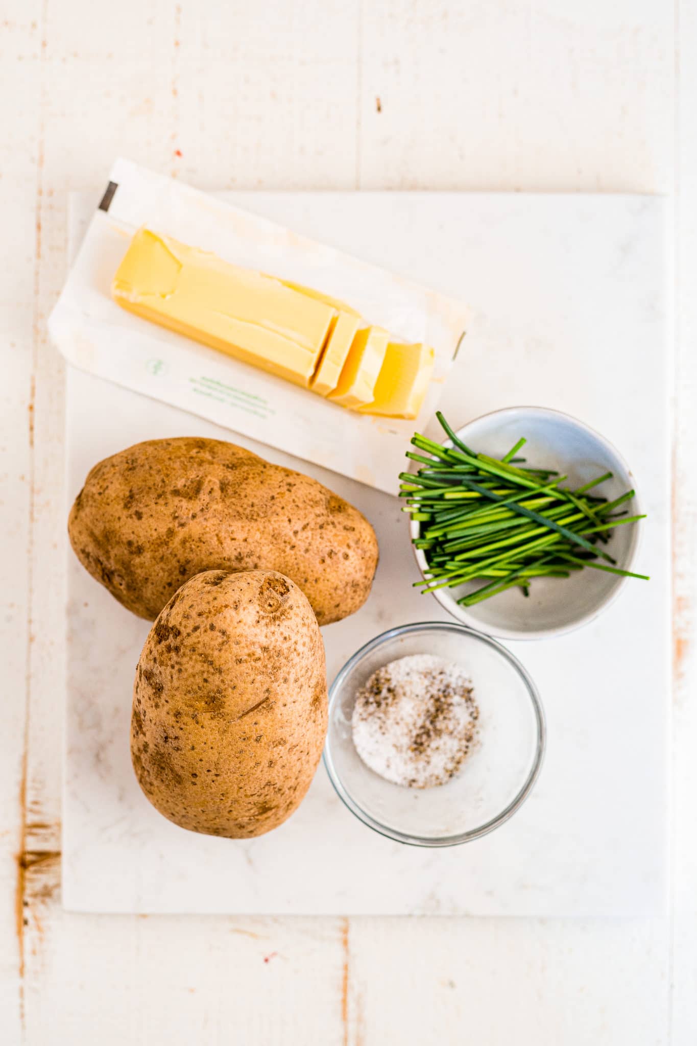 white cutting board with russet potatoes, salt, pepper, chives and butter