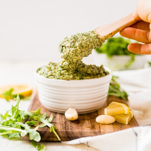 wooden spoon scooping out a serving of fresh arugula pesto from a white serving bowl on a wooden cutting board