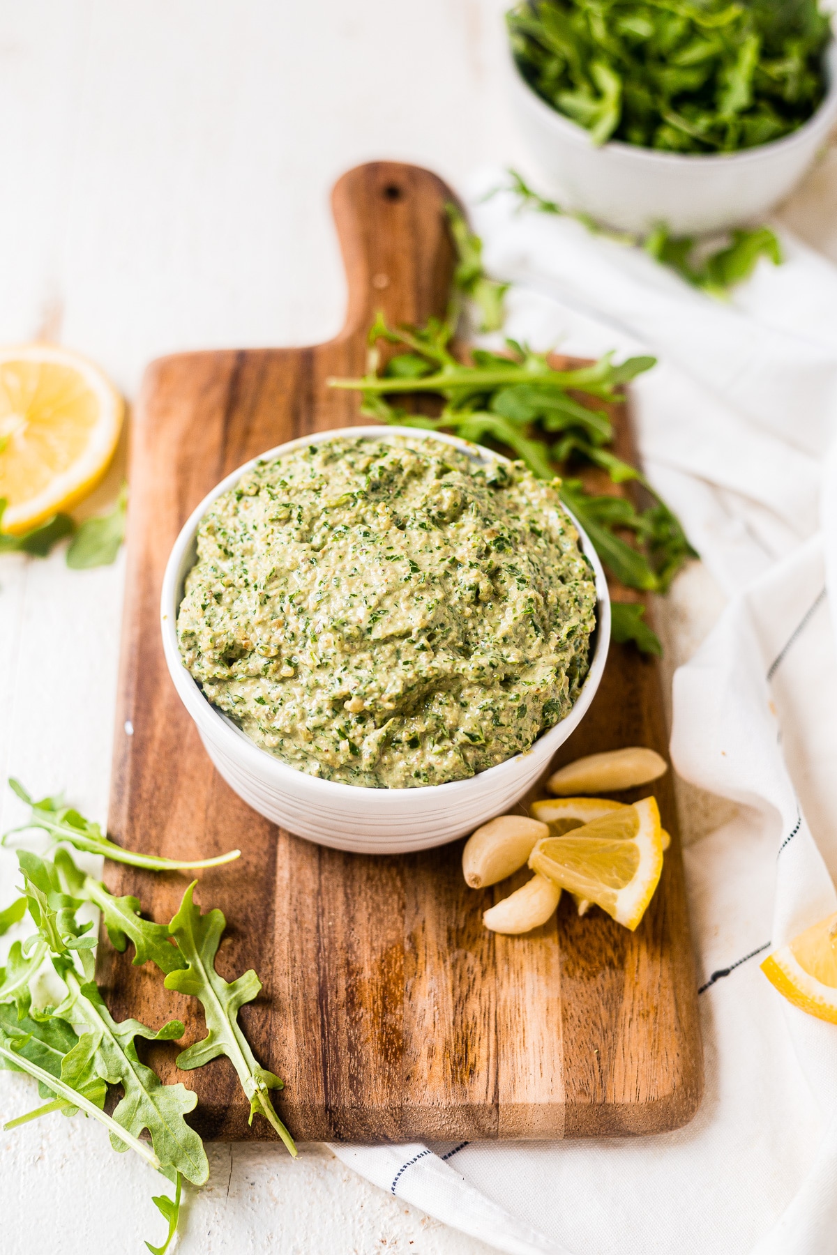 arugula pesto in a white serving dish on a cutting board surrounded by fresh lemon