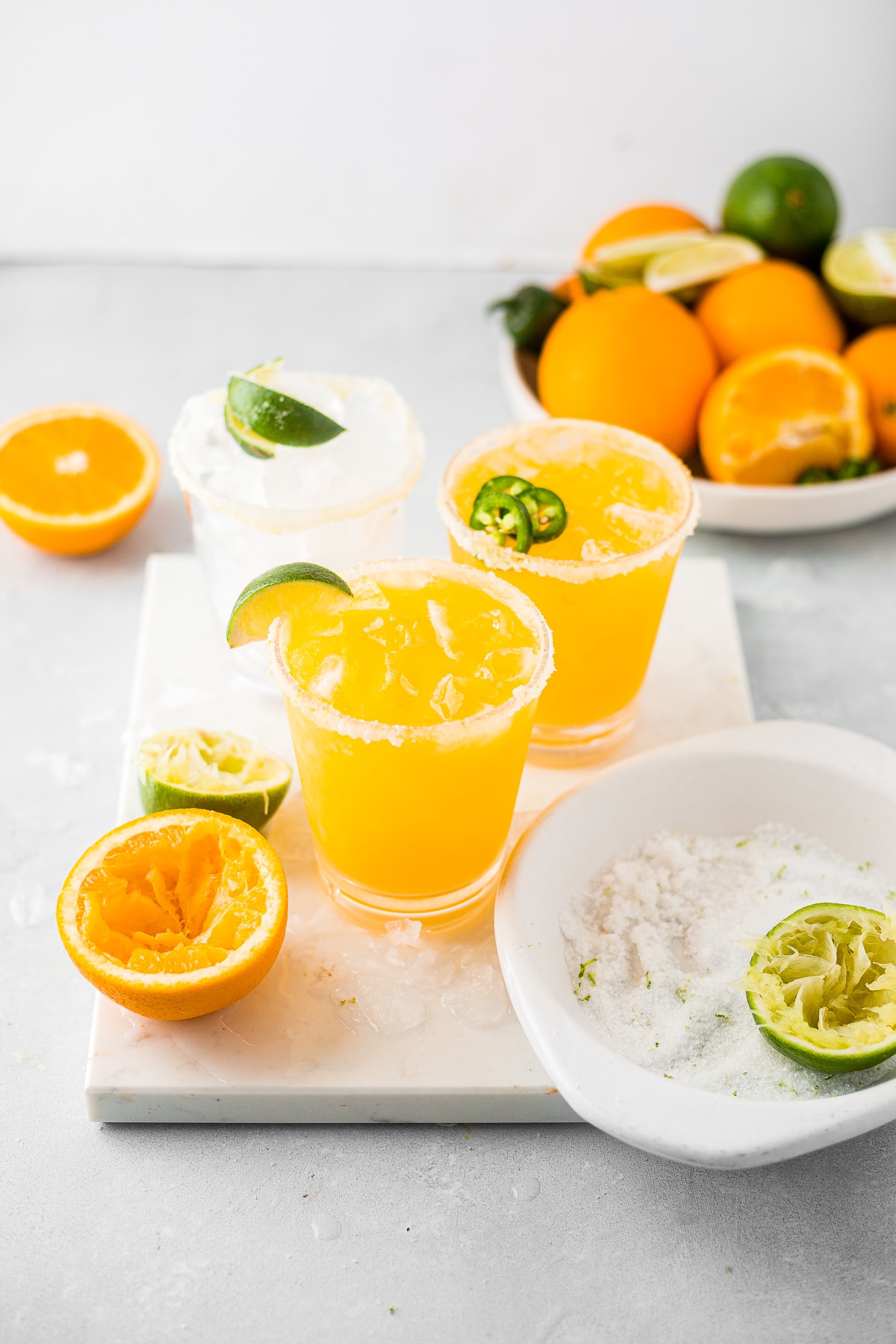 two mango margaritas on a marble slab garnished with salt and limes and surrounded by fresh oranges