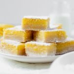 7 gluten free lemon bars stacked on a white plate so you can see the layers of shortbread crust and lemon curd then topped with confectioners' sugar