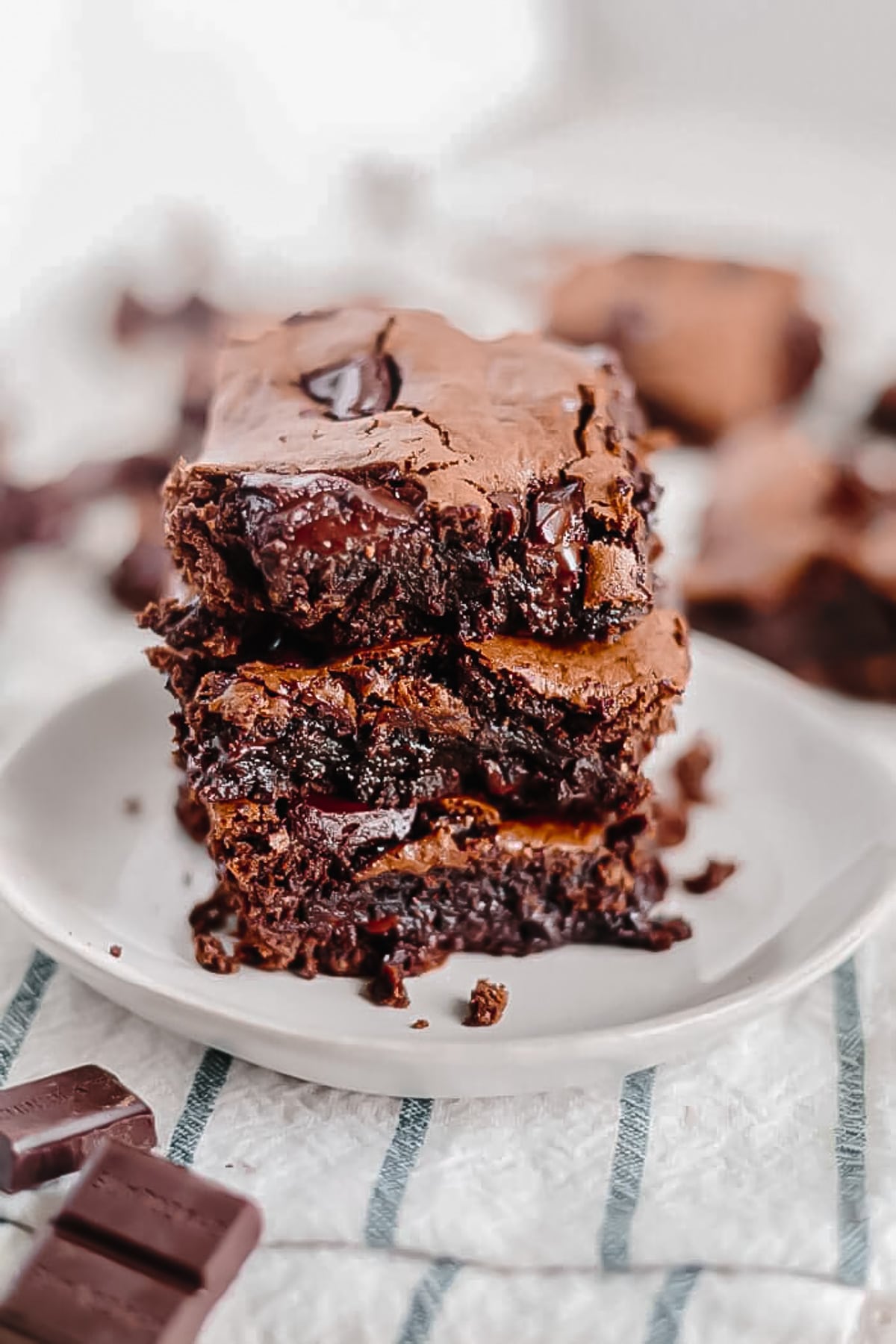 a stack of three paleo brownies on top of each other on a white plate with a blue striped napkin and chocolate chunks next to it