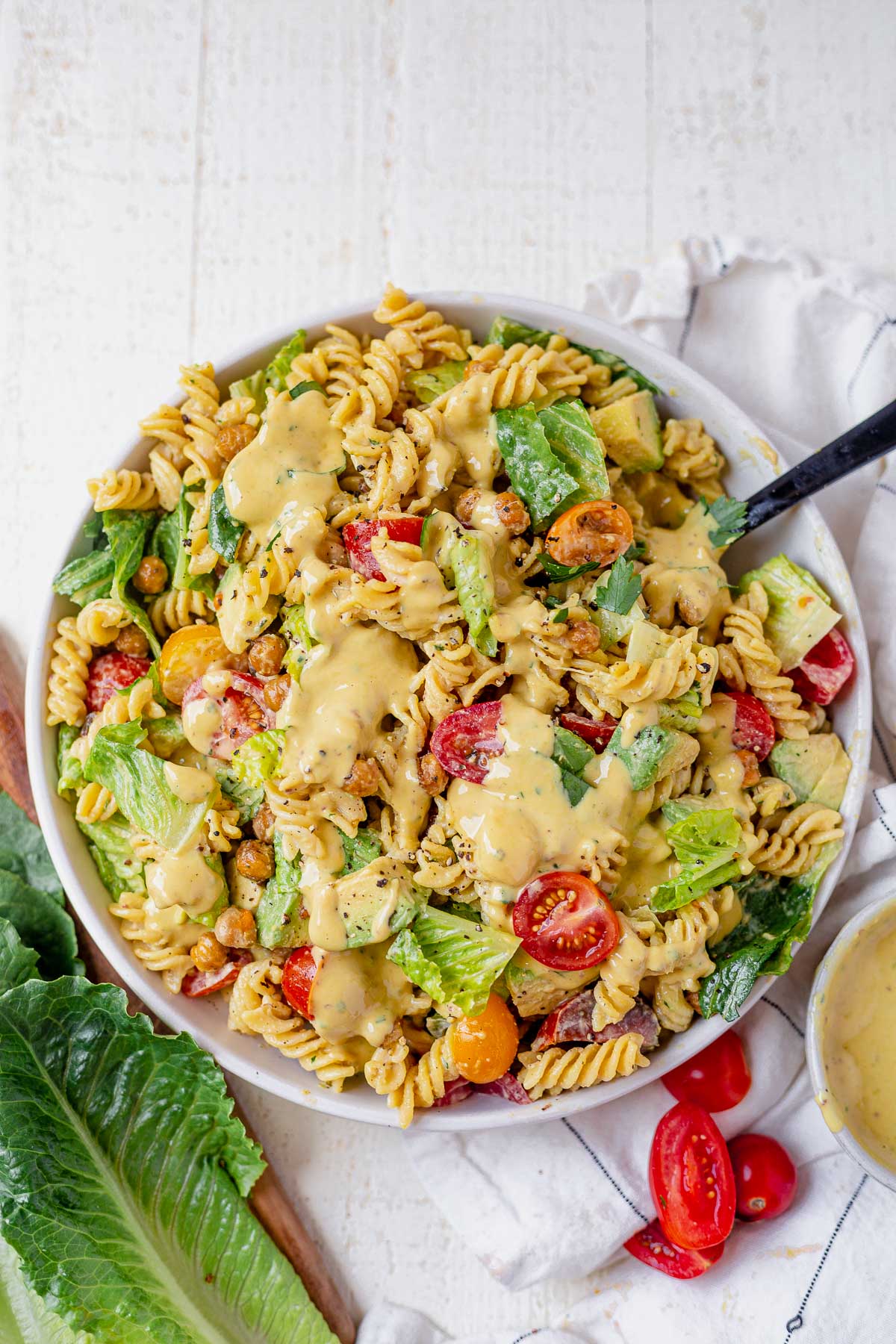 creamy caesar pasta salad in a big white serving bowl topped with more caesar dressing and romaine and tomatoes surrounding it