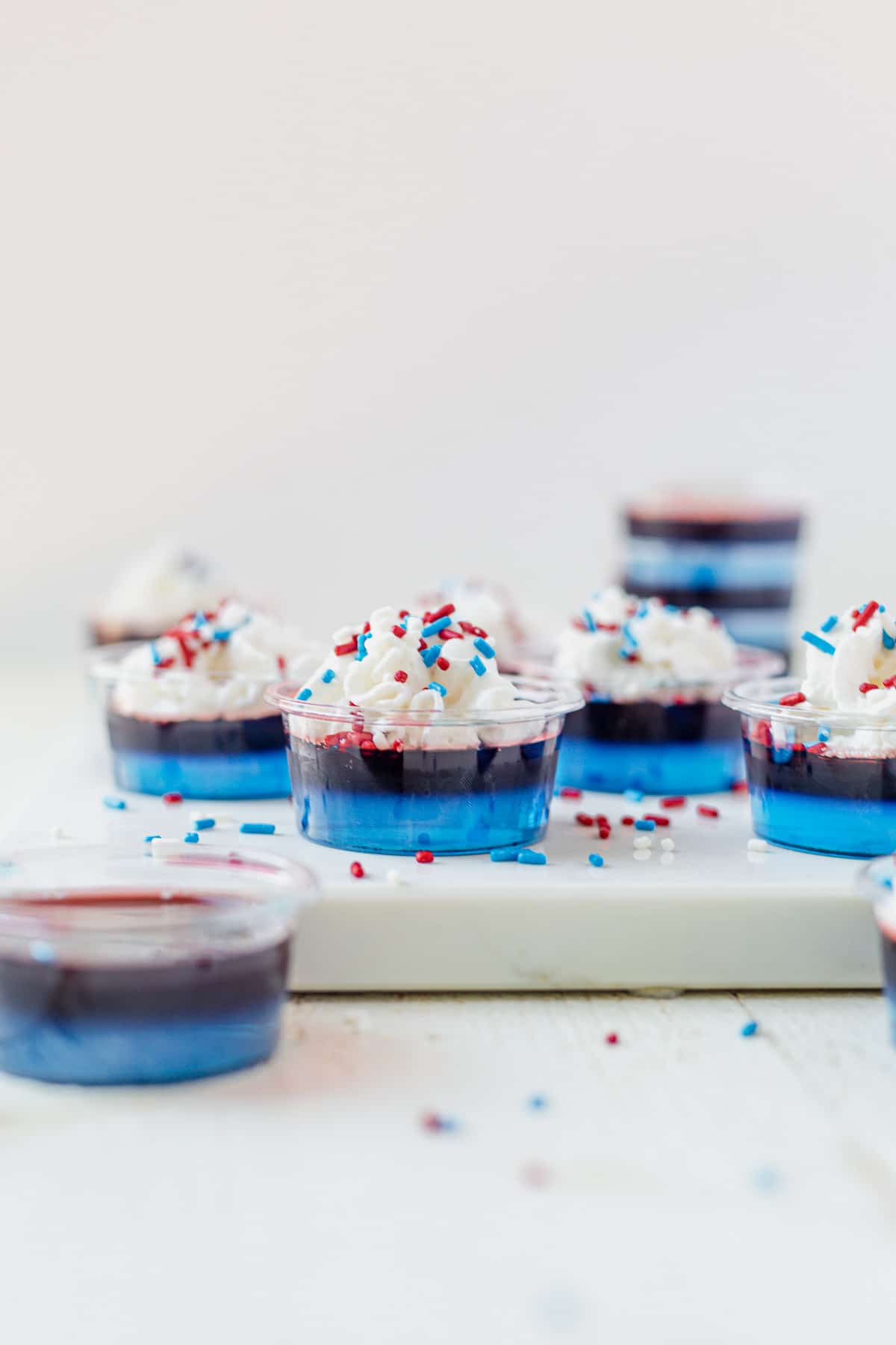 jello shots layered with blue and red jello then topped with whipped cream and sprinkles on a white serving tray