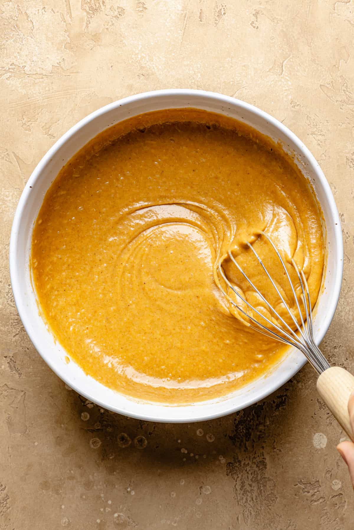 A hand is whisking smooth, orange pumpkin cobbler batter in a white mixing bowl on a textured beige surface, preparing the base for a cozy snickerdoodle cobbler.