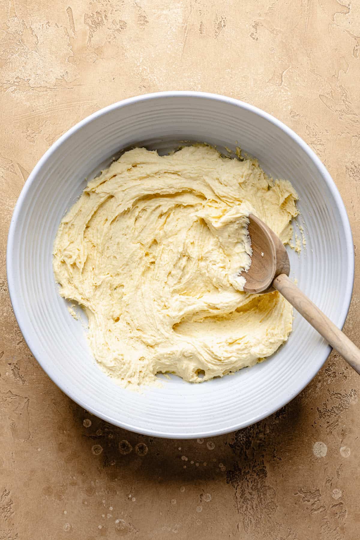 A white bowl with creamy yellow snickerdoodle cobbler cake batter being mixed with a wooden spoon, set on a light brown textured surface.