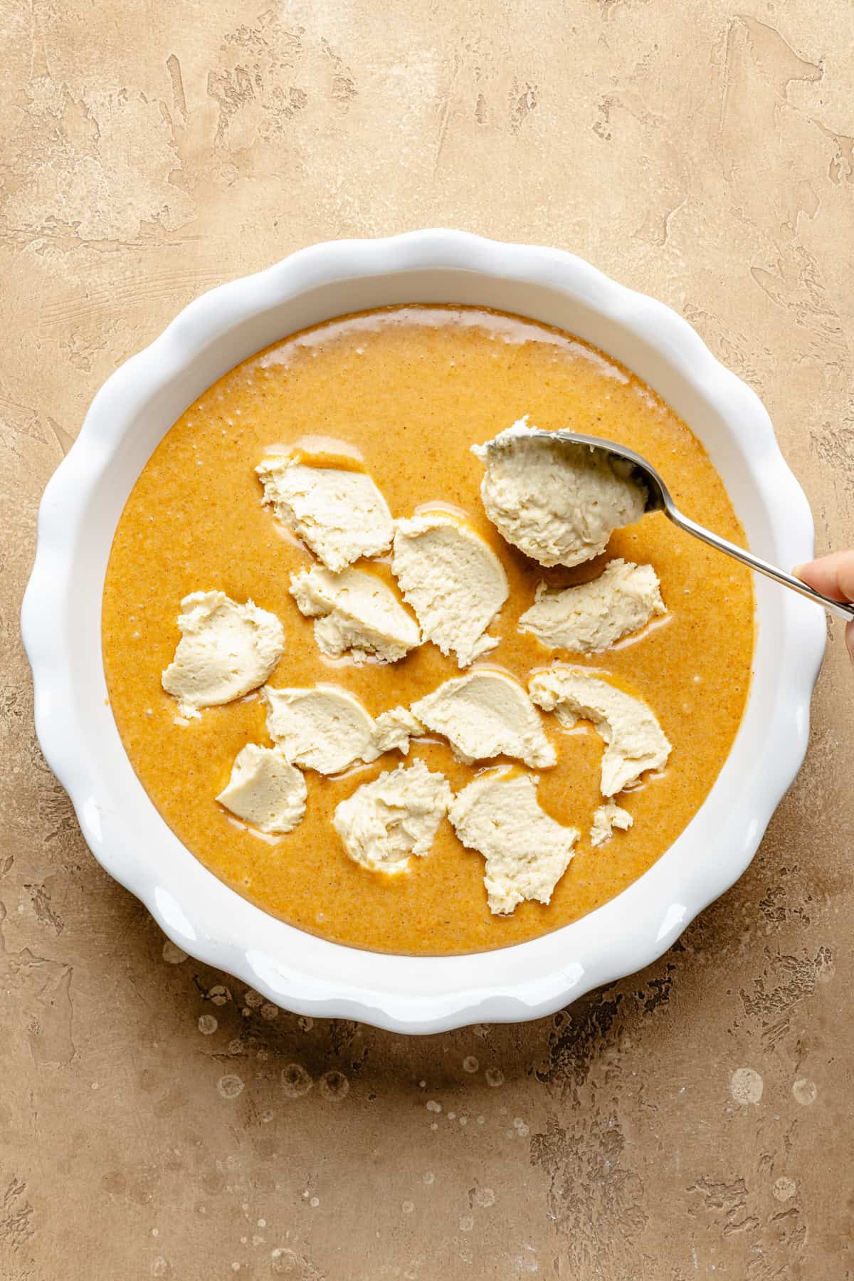 A hand holds a spoon, adding dollops of cobbler topping to a dish filled with pumpkin batter, on a light brown textured surface.