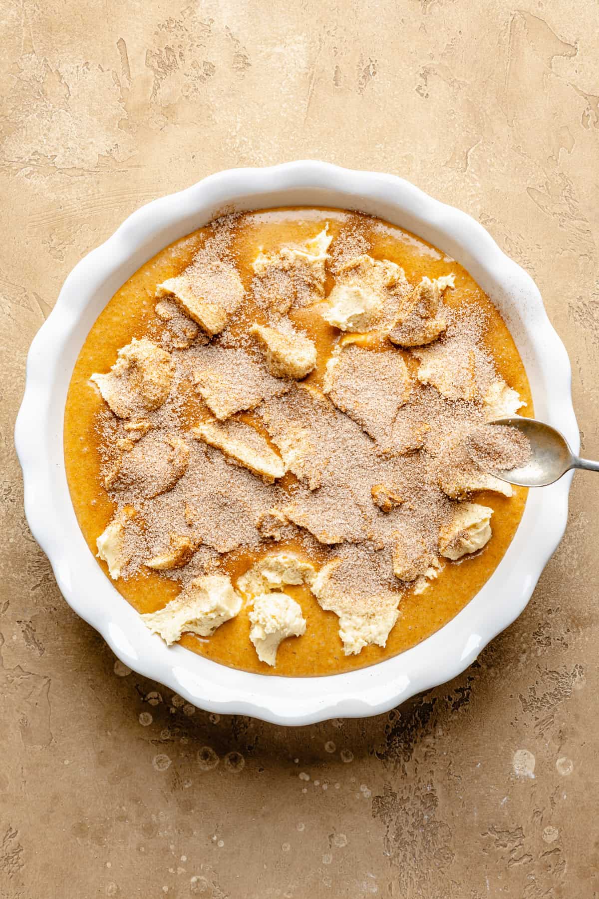 A round baking dish filled with an orange snickerdoodle pumpkin cobbler mixture, sprinkled with cinnamon sugar. A spoon rests on the right edge of the dish.