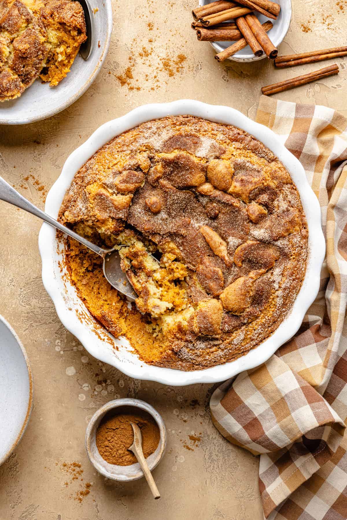 A snickerdoodle pumpkin cobbler with a golden, cinnamon-topped crust sits baked to perfection in a white dish. A spoon has scooped out a portion, revealing the warm, gooey center. Nearby are cinnamon sticks, a bowl of ground cinnamon, a checked cloth, and empty plates.