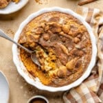 Baked snickerdoodle cobbler with a golden cinnamon sugar crust in a white round dish, with a serving spoon taking out a portion. A checkered napkin and plates are nearby on a beige surface.
