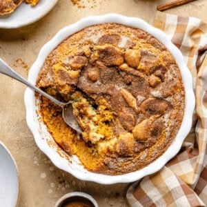 Baked snickerdoodle cobbler with a golden cinnamon sugar crust in a white round dish, with a serving spoon taking out a portion. A checkered napkin and plates are nearby on a beige surface.