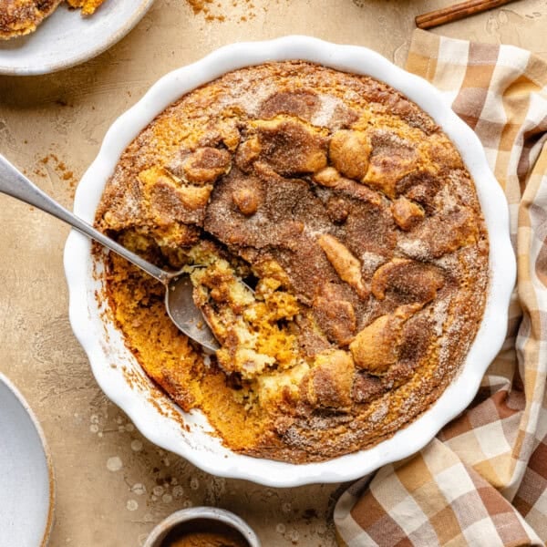 Baked snickerdoodle cobbler with a golden cinnamon sugar crust in a white round dish, with a serving spoon taking out a portion. A checkered napkin and plates are nearby on a beige surface.
