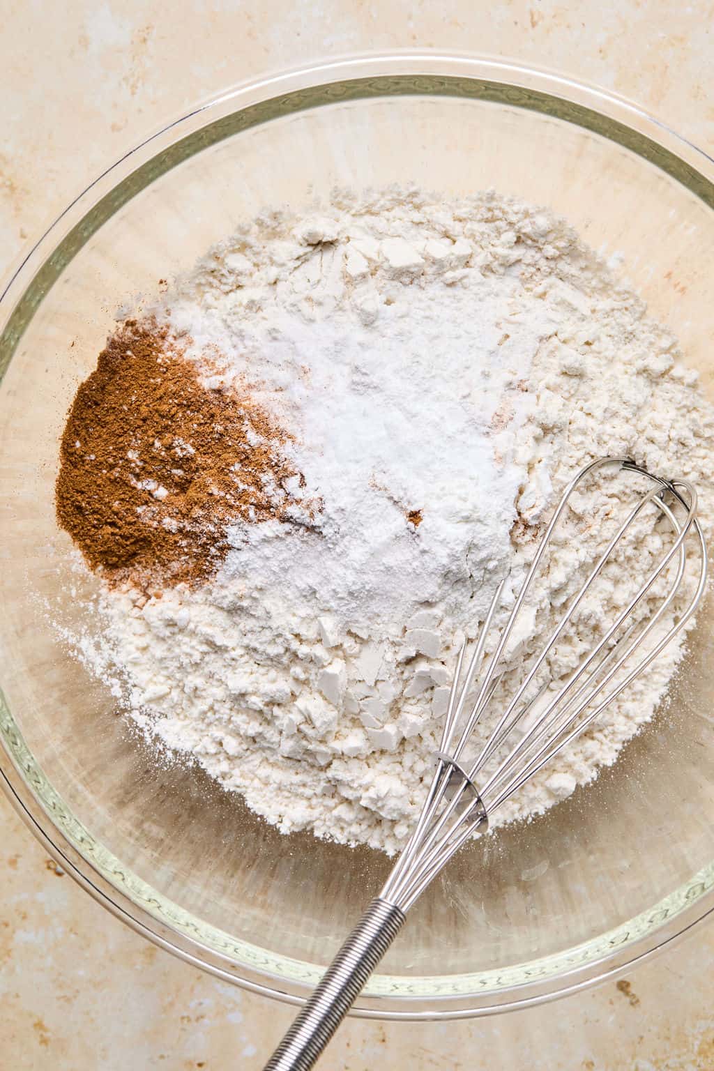 A glass bowl filled with flour, baking powder, and ground cinnamon for gluten-free pumpkin muffins, with a metal whisk resting on the right side of the bowl, ready for mixing.