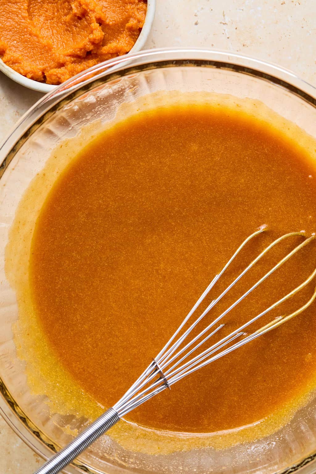 A glass bowl filled with pumpkin pie filling mixture and a metal whisk—perfect for making gluten-free pumpkin muffins. In the upper left corner, a small bowl holds vibrant orange pumpkin puree.