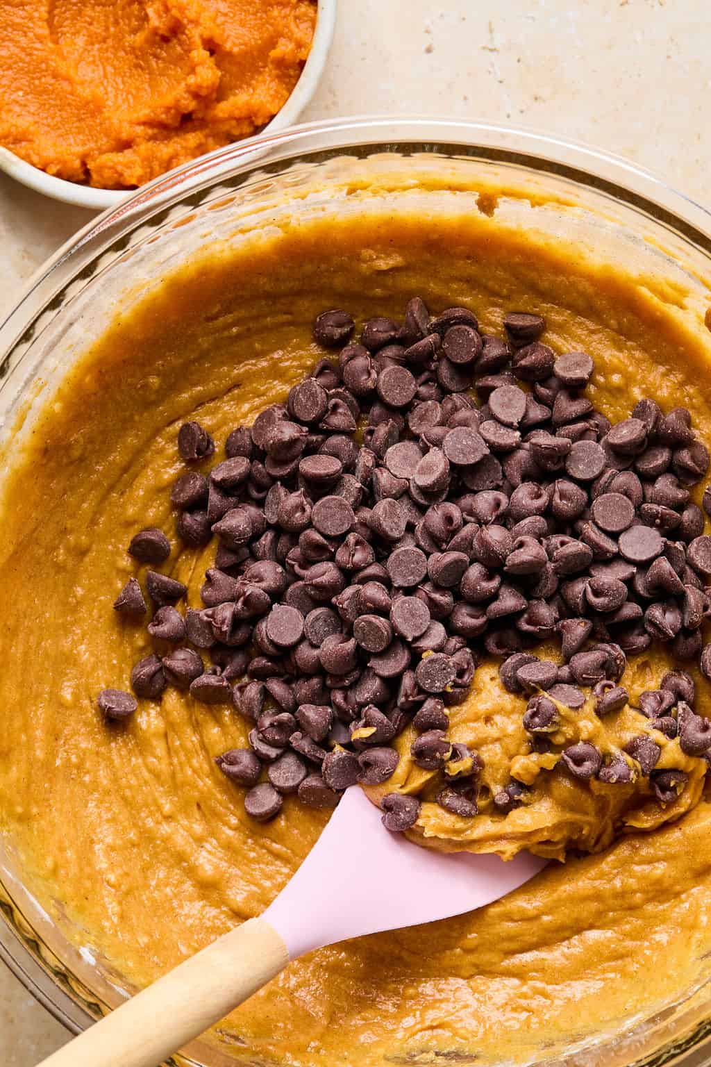 A mixing bowl of pumpkin batter for gluten-free pumpkin muffins with chocolate chips being stirred in using a pink spatula. A small bowl of pumpkin puree sits in the background.
